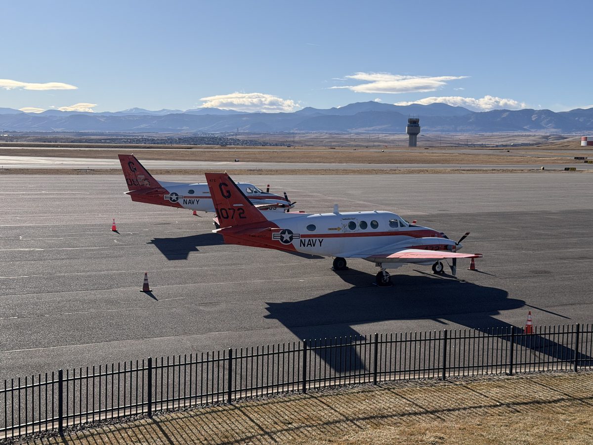 Two U.S. Navy T-44C Pegasus trainers in red and white livery parked on the ramp at Rocky Mountain Metropolitan Airport with the control tower and snow-capped Rocky Mountains in the background
